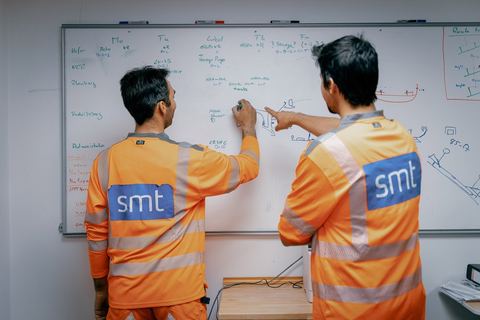 To SMT-medarbejdere gennemgår en moniteringsopgave ved en whiteboard-tavle